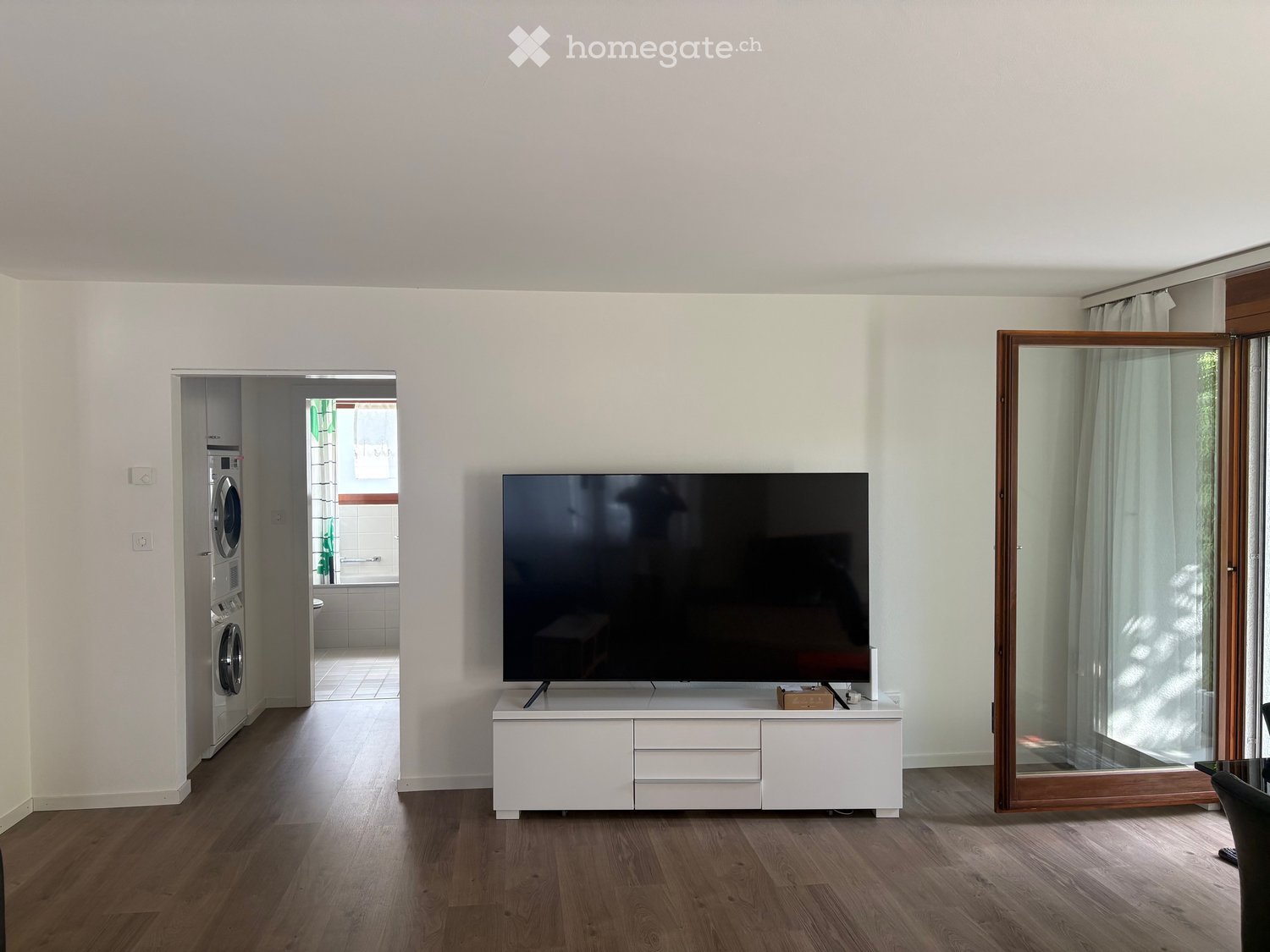 Modern living room with hardwood flooring, large flat-screen TV on a white stand, and a private washing machine in the adjacent space.