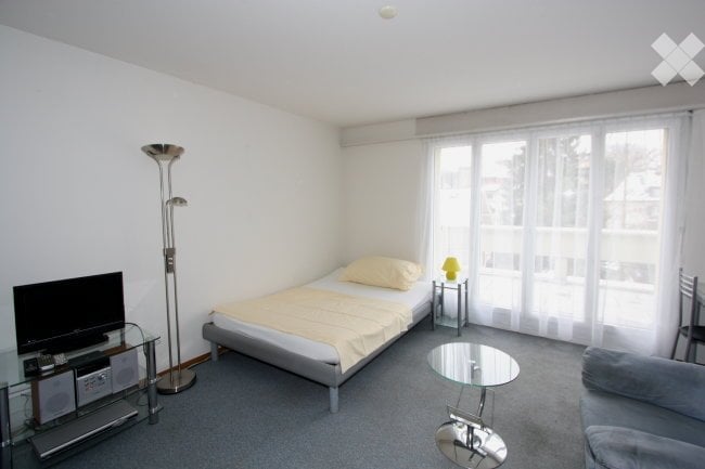 Cozy bedroom with a single bed, nightstand, table lamp, chair, sofa, television, and floor lamp. Large windows with white curtains let in natural light.