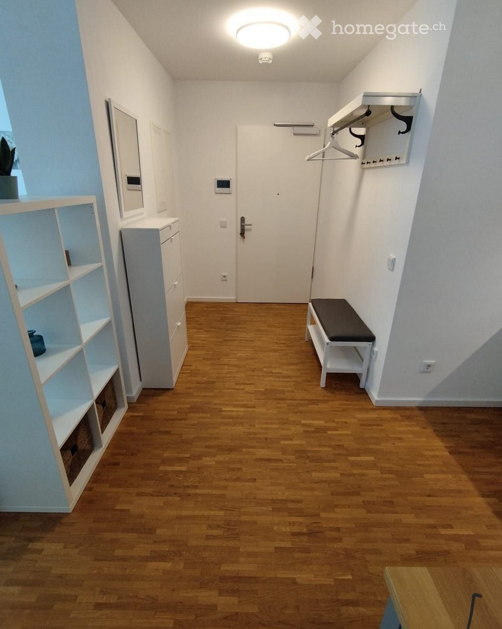 A narrow hallway with wooden flooring, white walls, and a door at the end. Features include a small bench, a coat rack, a shelf with decorative items, and a mirror on the wall.