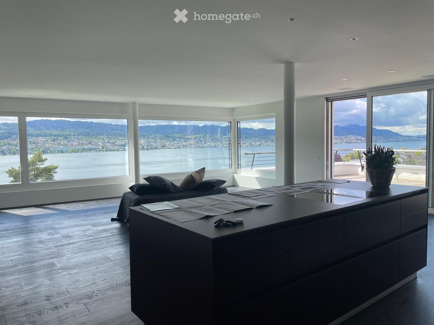 Modern, open-concept living space with large windows offering a scenic view of a lake and mountains. Features a dark wooden coffee table, a sofa with pillows, and a potted plant on the balcony.