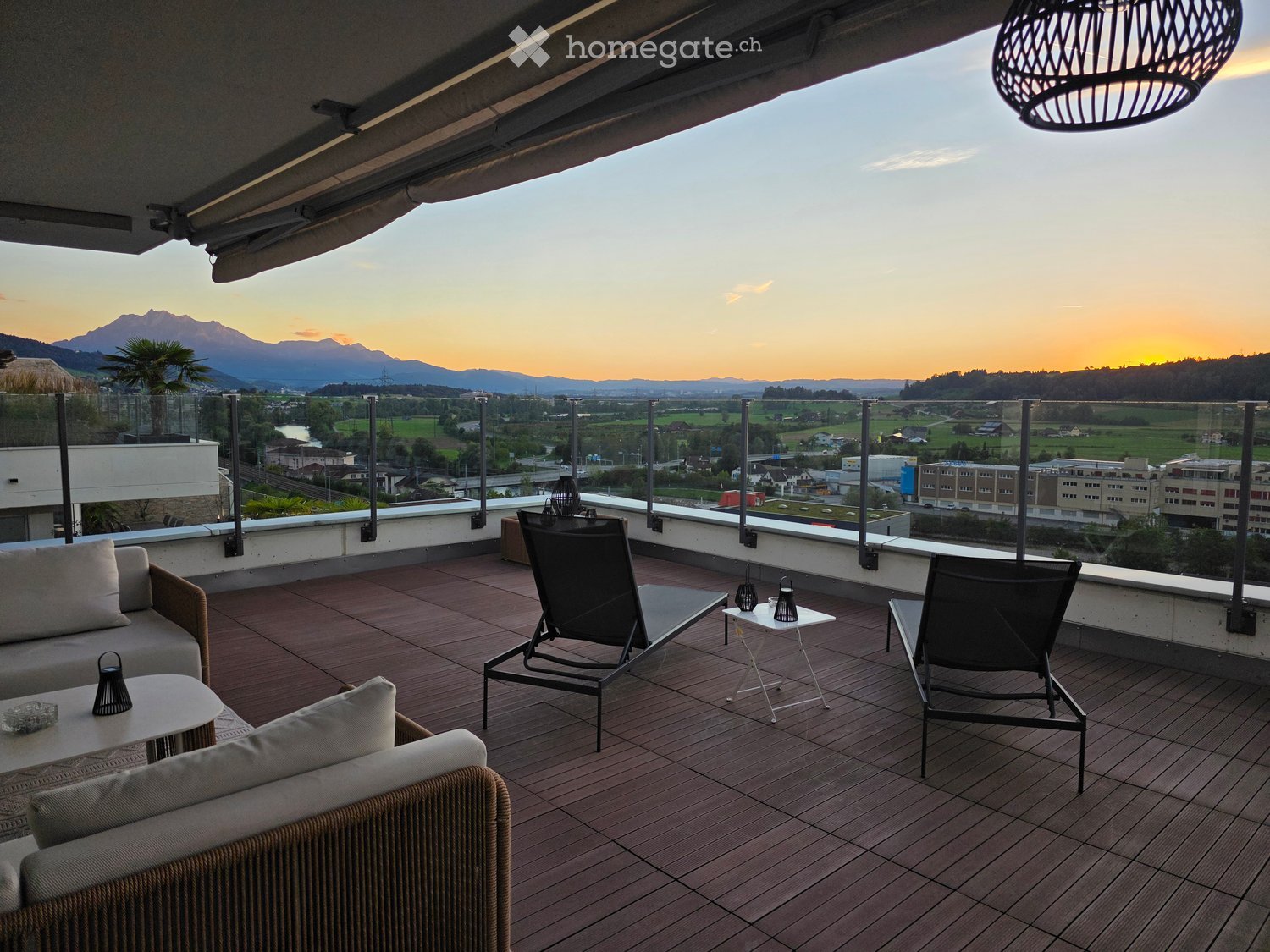 Balcony with wooden decking, sofa, chairs, small tables, and a scenic mountain view at sunset.