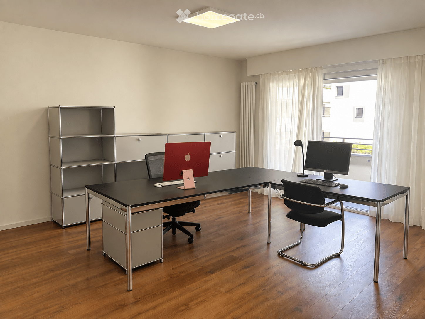 Modern office space with wooden flooring, large window with white curtains, two desks with chairs, laptops, and a shelf unit.