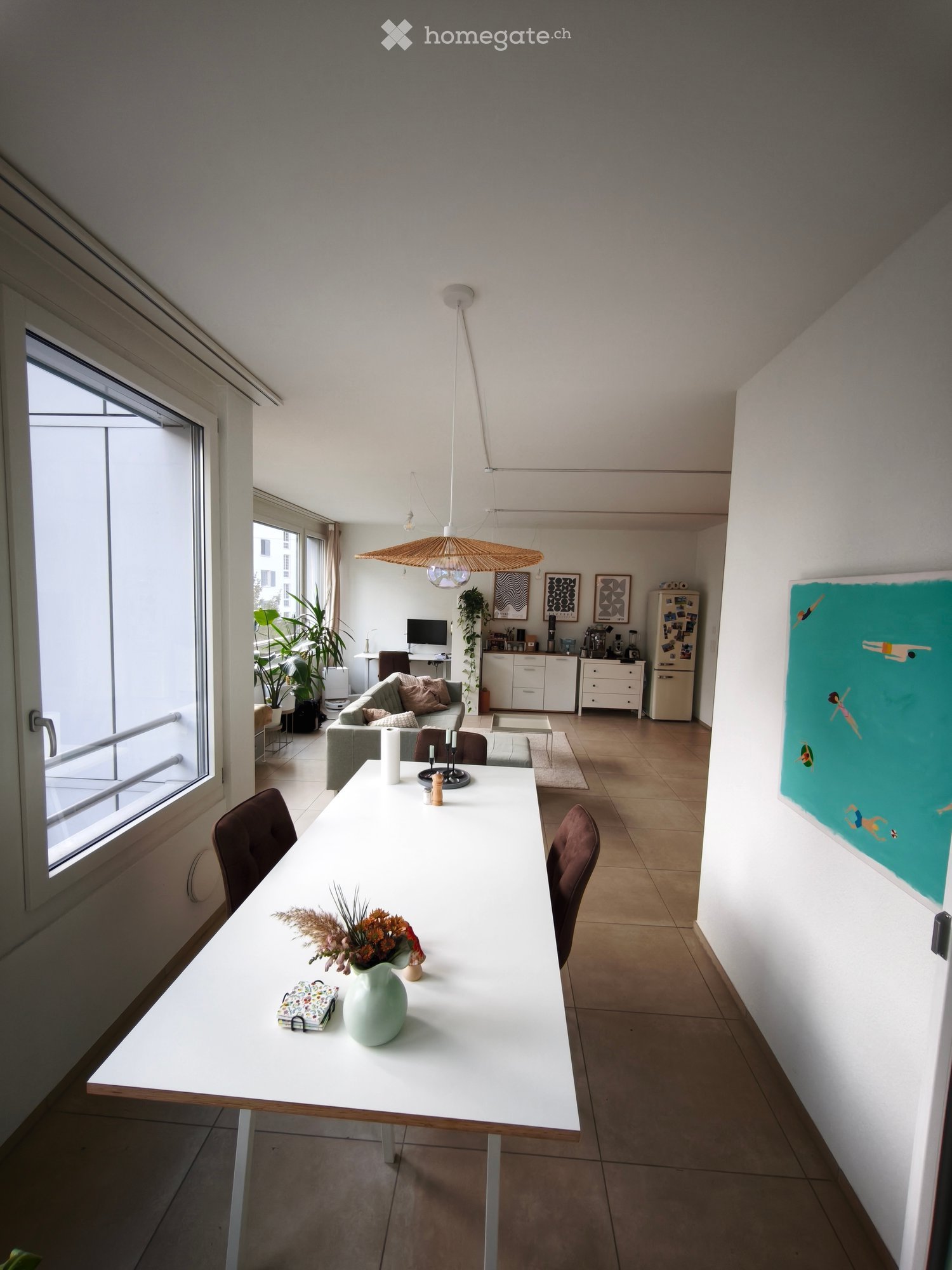 Open-plan living and dining area with a white dining table, chairs, a grey sofa, a refrigerator, and a large window with a view.