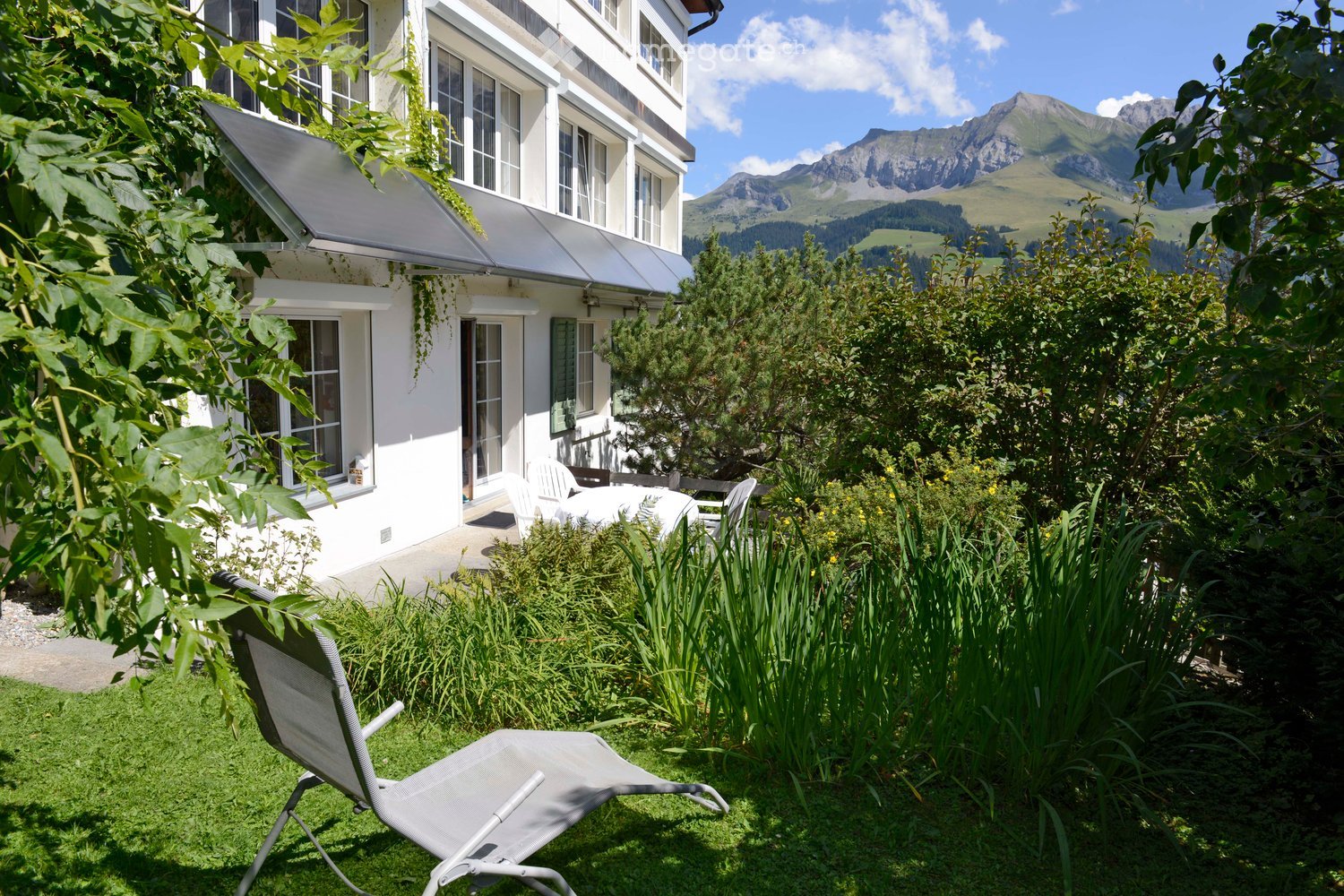A well-maintained garden area with a lounge chair, surrounded by lush greenery and a view of a multi-story white building with large windows and a covered patio. The background features a scenic mountain range under a clear blue sky.