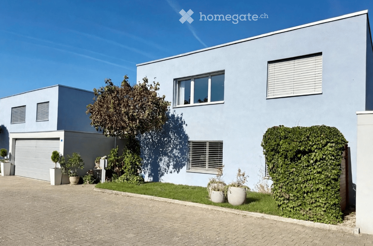 Modern two-story house with light blue exterior, garage, and small garden area with potted plants.