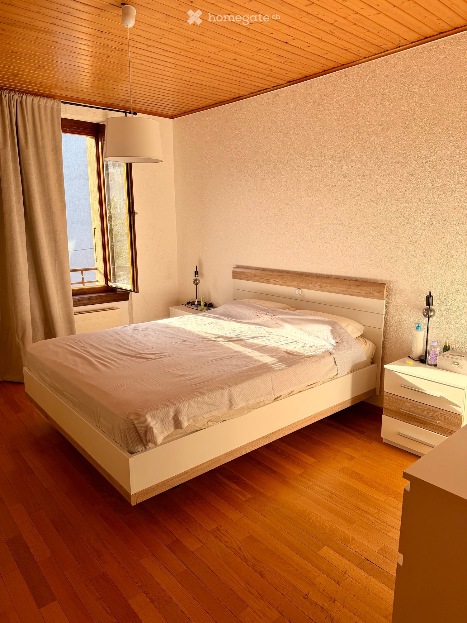 Cozy bedroom with wooden floor, large bed with white bedding, nightstand with lamp, and a window with curtains.