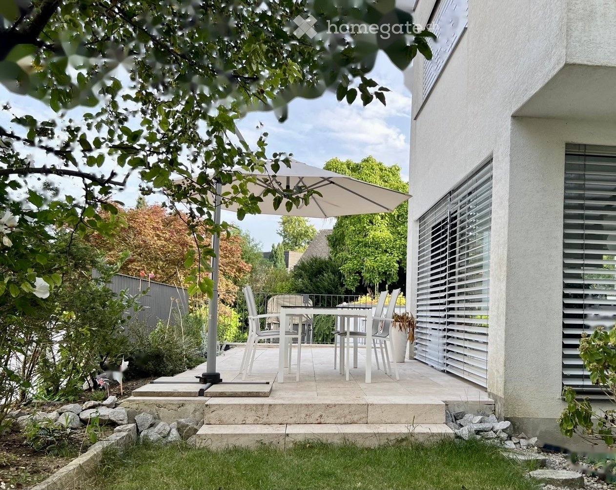 A modern outdoor patio area with a white dining set, umbrella, and surrounding greenery.