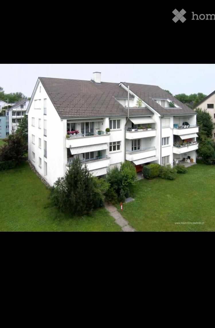A multi-story residential building with multiple balconies, surrounded by greenery and a well-maintained lawn.