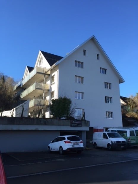 Multi-story residential building with white exterior, multiple balconies, and a parking area with several cars.