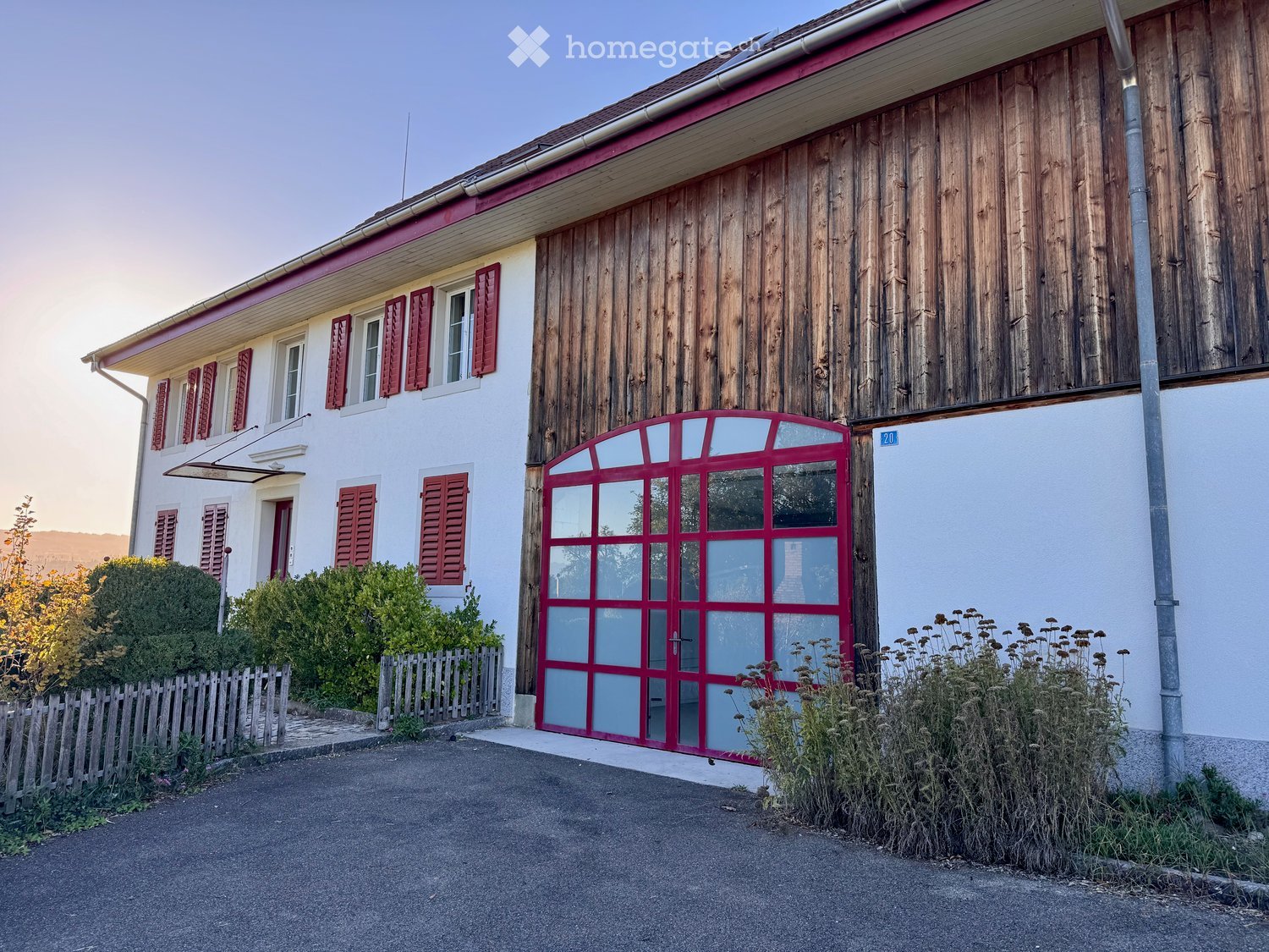 Two-story house with a red garage door, white walls, and red shutters on the windows. The house has a small garden area with a wooden fence and some bushes.
