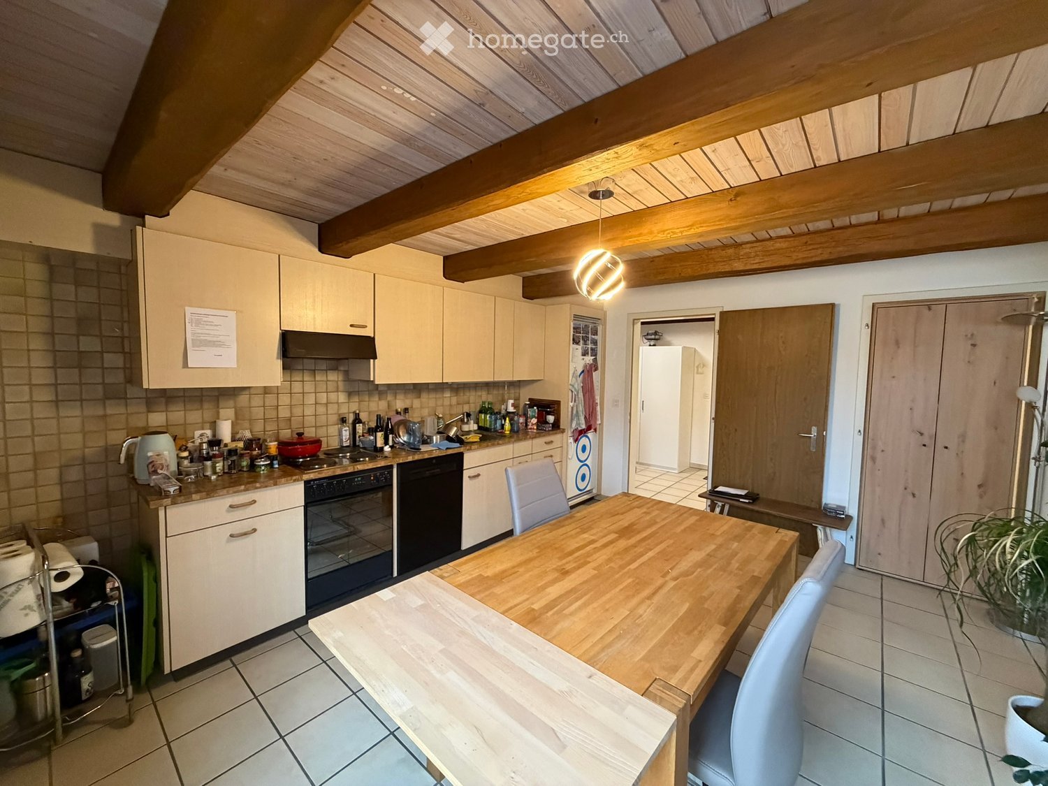 Bright kitchen with light wood cabinets, a central wooden dining table, and modern appliances including a stove, dishwasher, and refrigerator. Exposed wooden beams on the ceiling and pendant lighting.