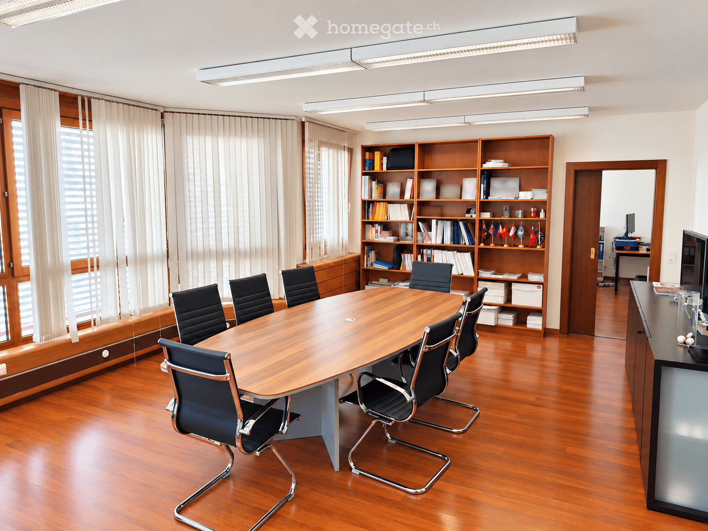 Conference room with a large wooden table, black chairs, bookshelf filled with books and flags, large windows with curtains, and a screen on a stand.