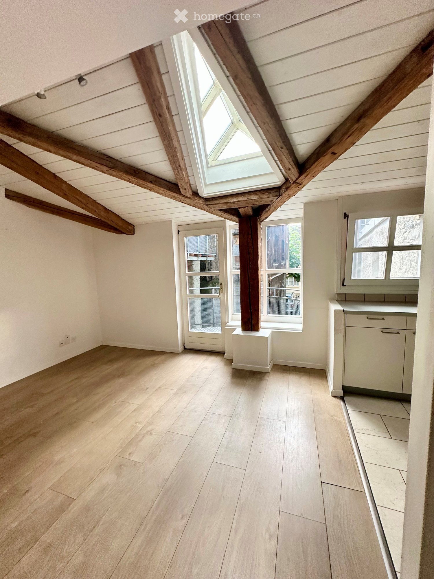 Empty attic space with wooden beams, skylight window, wooden flooring, and white walls. No furniture or equipment present.