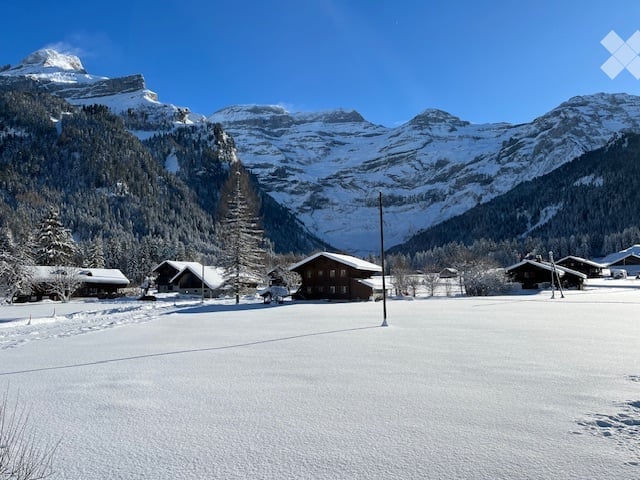 Snow-covered field with traditional wooden chalets and snow-capped mountains in the background.