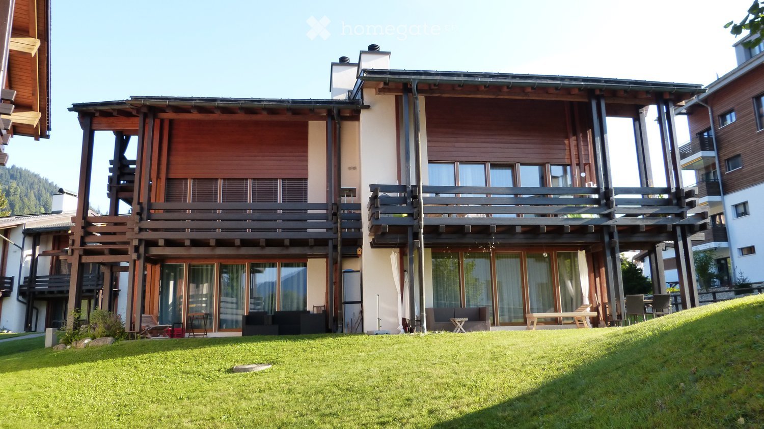Modern multi-story building with wooden balconies and large windows, surrounded by a well-maintained lawn.