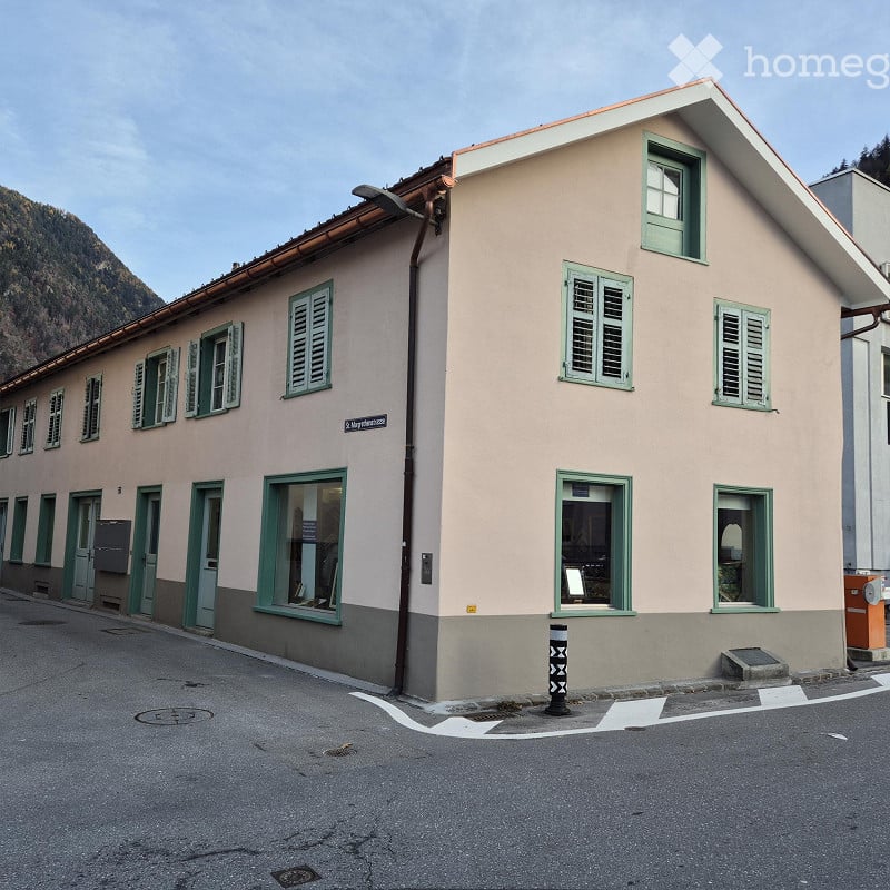 Two-story building with light pink facade, green shutters, and multiple windows. The building has a sloped roof and is situated on a street with a visible mountain in the background.