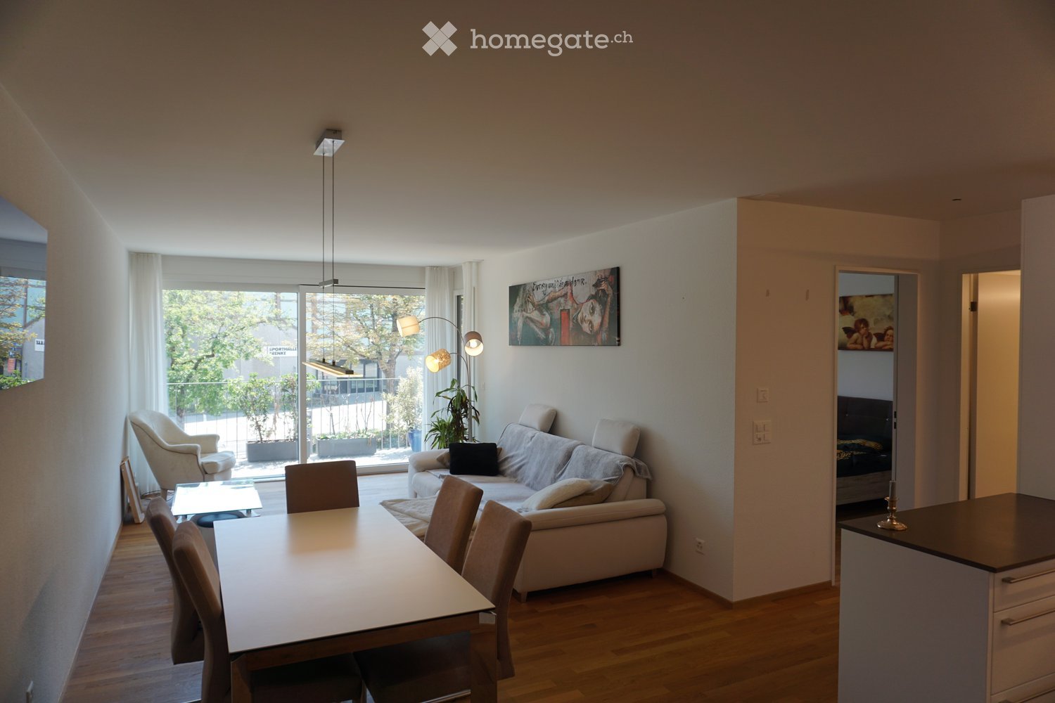 Modern living and dining area with a sofa, dining table, chairs, armchair, floor lamp, table lamp, and artwork on the wall. The space features large windows with sheer curtains and a view of a balcony area with plants.