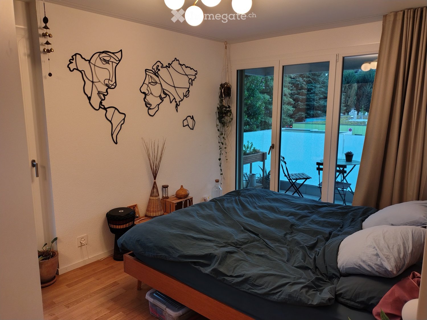 Cozy bedroom with a wooden bed, nightstand, small table, and decorative wall art. Large sliding glass doors lead to a balcony with outdoor furniture.