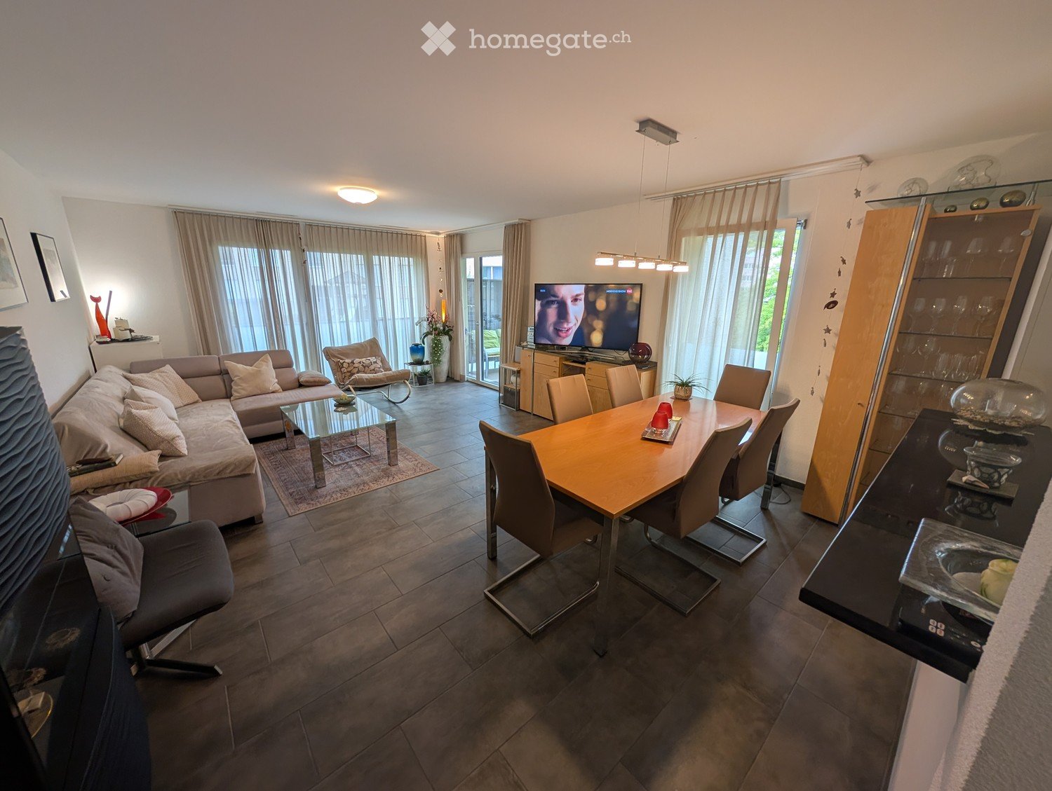 Modern living and dining area with a grey sectional sofa, glass coffee table, dining table with chairs, television, and decorative items.