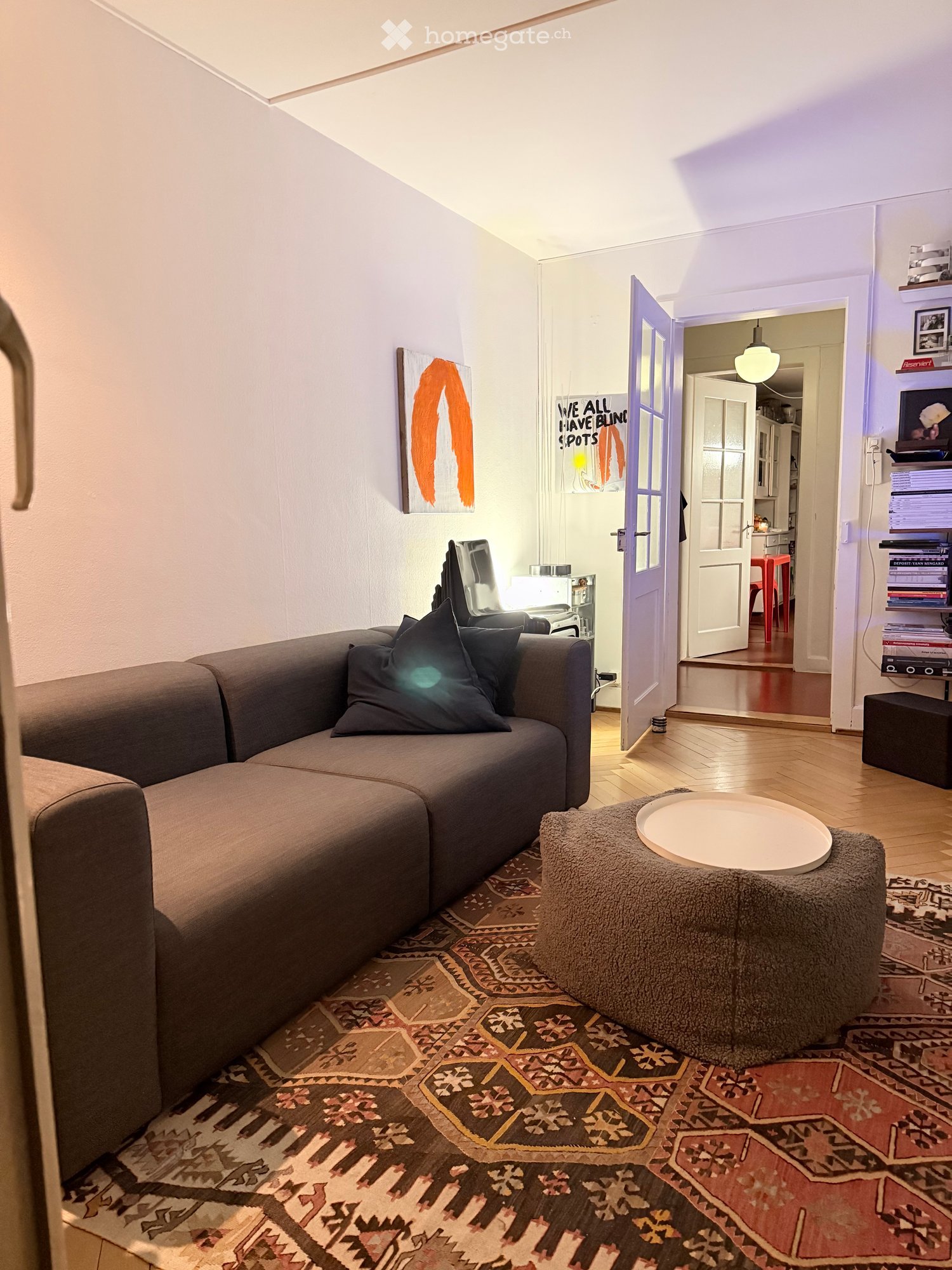 Living room with a grey sectional sofa, ottoman, round table, shelves, and artwork on the wall. The room features wooden flooring and a patterned rug.