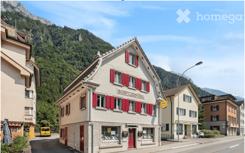Traditional Swiss building with red shutters and signage reading "Schwyzenseeli" in a picturesque village setting with mountains in the background.