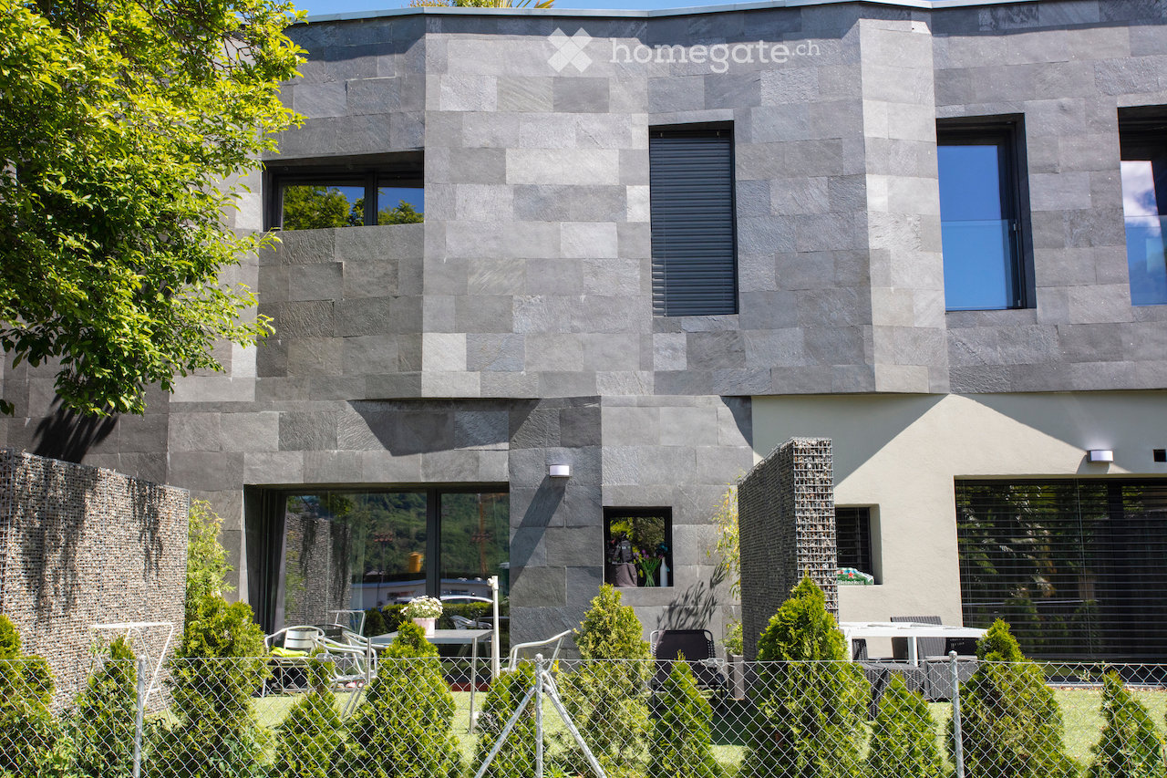 Modern two-story house with grey stone facade, large windows, and a small outdoor seating area with tables and chairs.