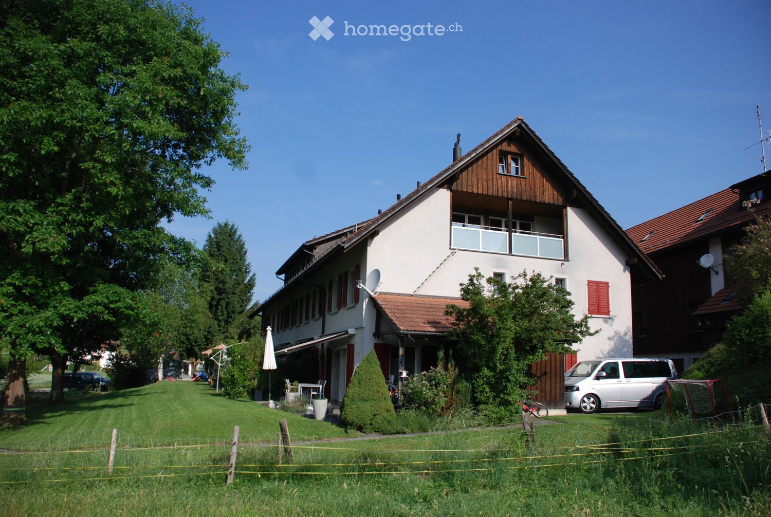 Two-story house with white walls, red shutters, and a balcony on the upper floor. The house has a red-tiled roof and is surrounded by a well-maintained lawn and trees.