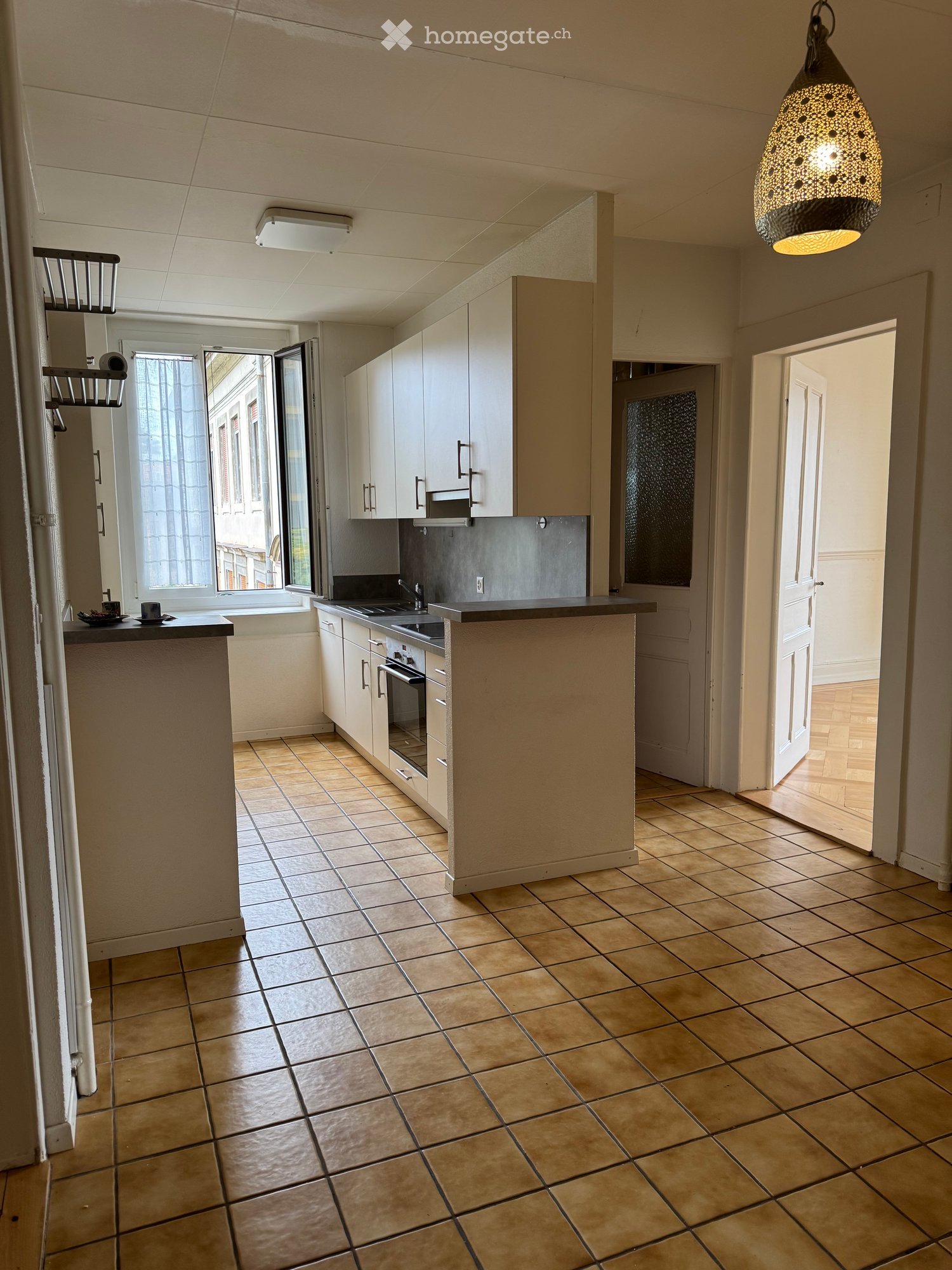 Modern kitchen with white cabinets, a central island, and a tiled floor. Features a sink, oven, and a hanging light fixture.