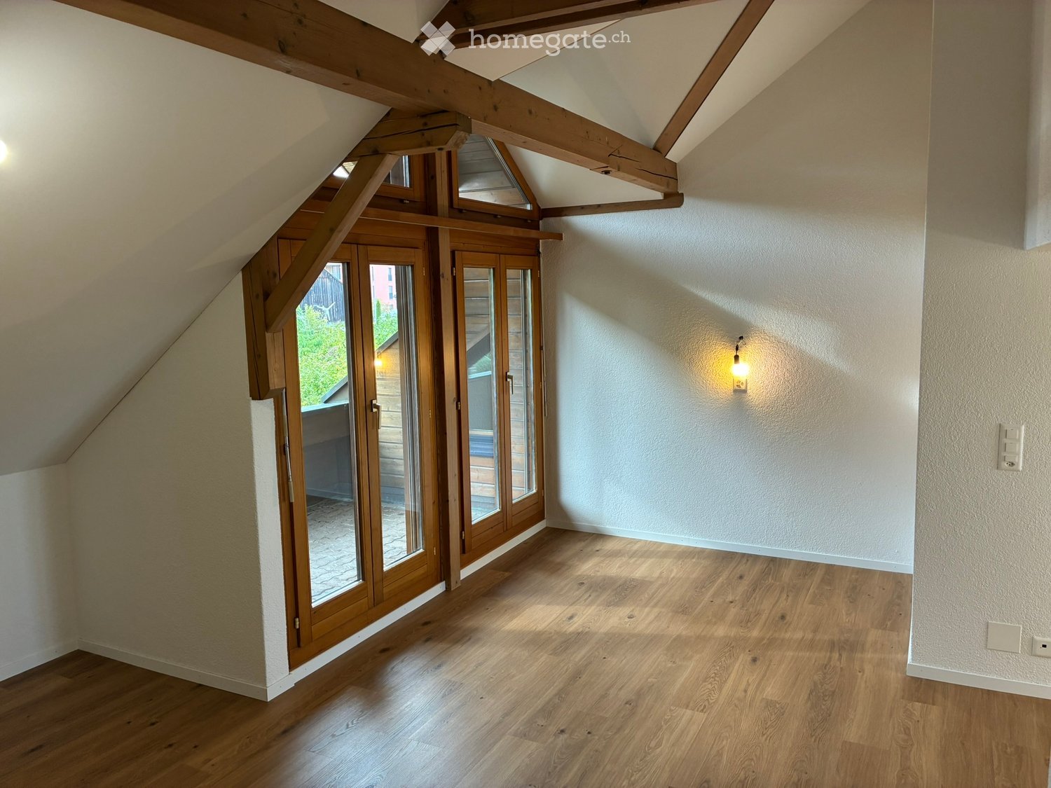 Empty attic room with wooden floors, exposed wooden beams, and large windows with wooden frames, allowing natural light to enter.