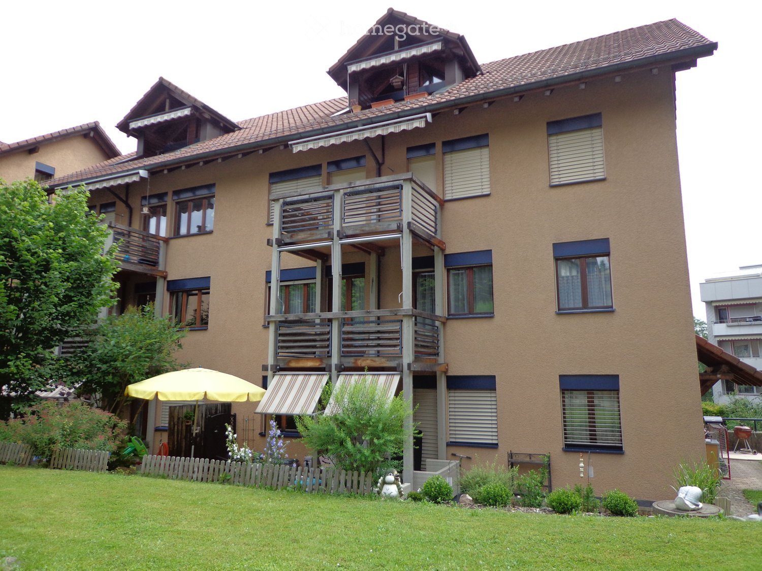 A two-story house with beige exterior walls, multiple windows, balconies, and a well-maintained garden with a yellow umbrella and small plants.
