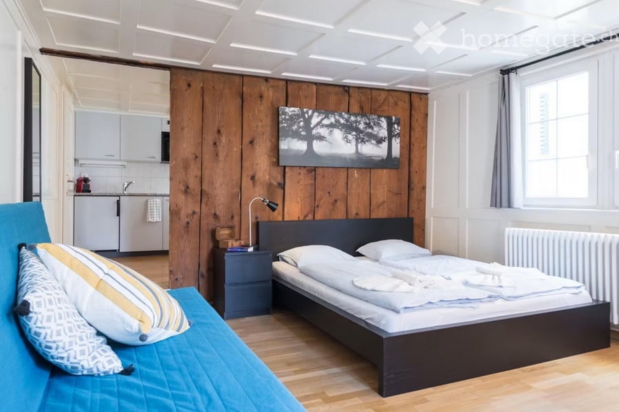 A modern, compact bedroom with a double bed, nightstand, sofa, and a small kitchen area with cabinets and a sink.