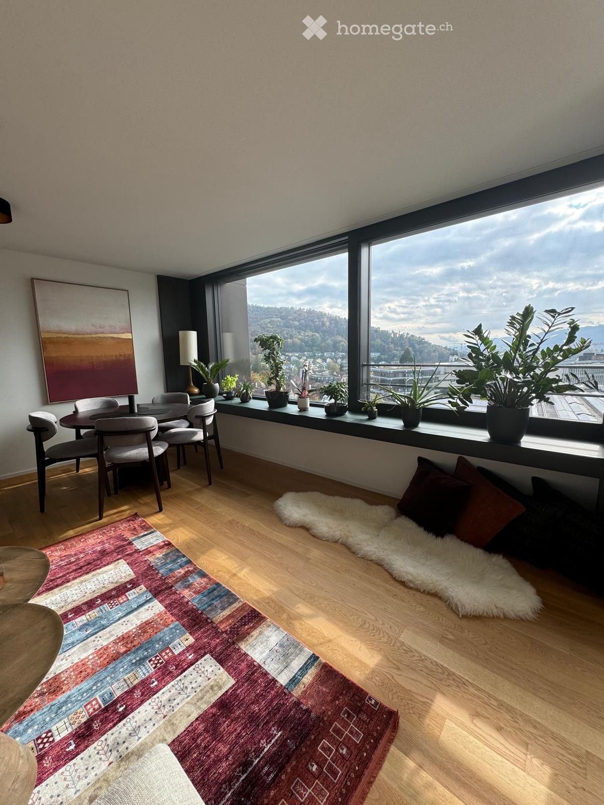 Bright living space with large windows offering a scenic view, featuring a dining area with a table and chairs, potted plants on the windowsill, a colorful rug, and a cozy seating area with throw pillows.