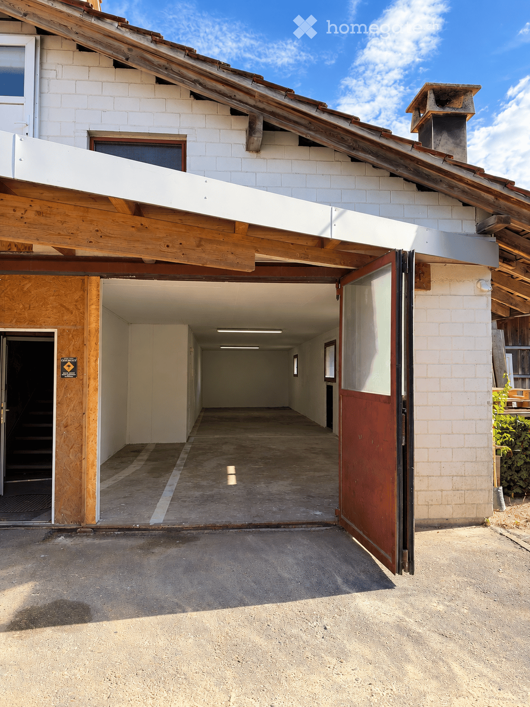 A modern building with a large garage entrance, white brick walls, wooden beams, and a slanted wooden roof. The garage has a concrete floor and is partially open, revealing a hallway inside.