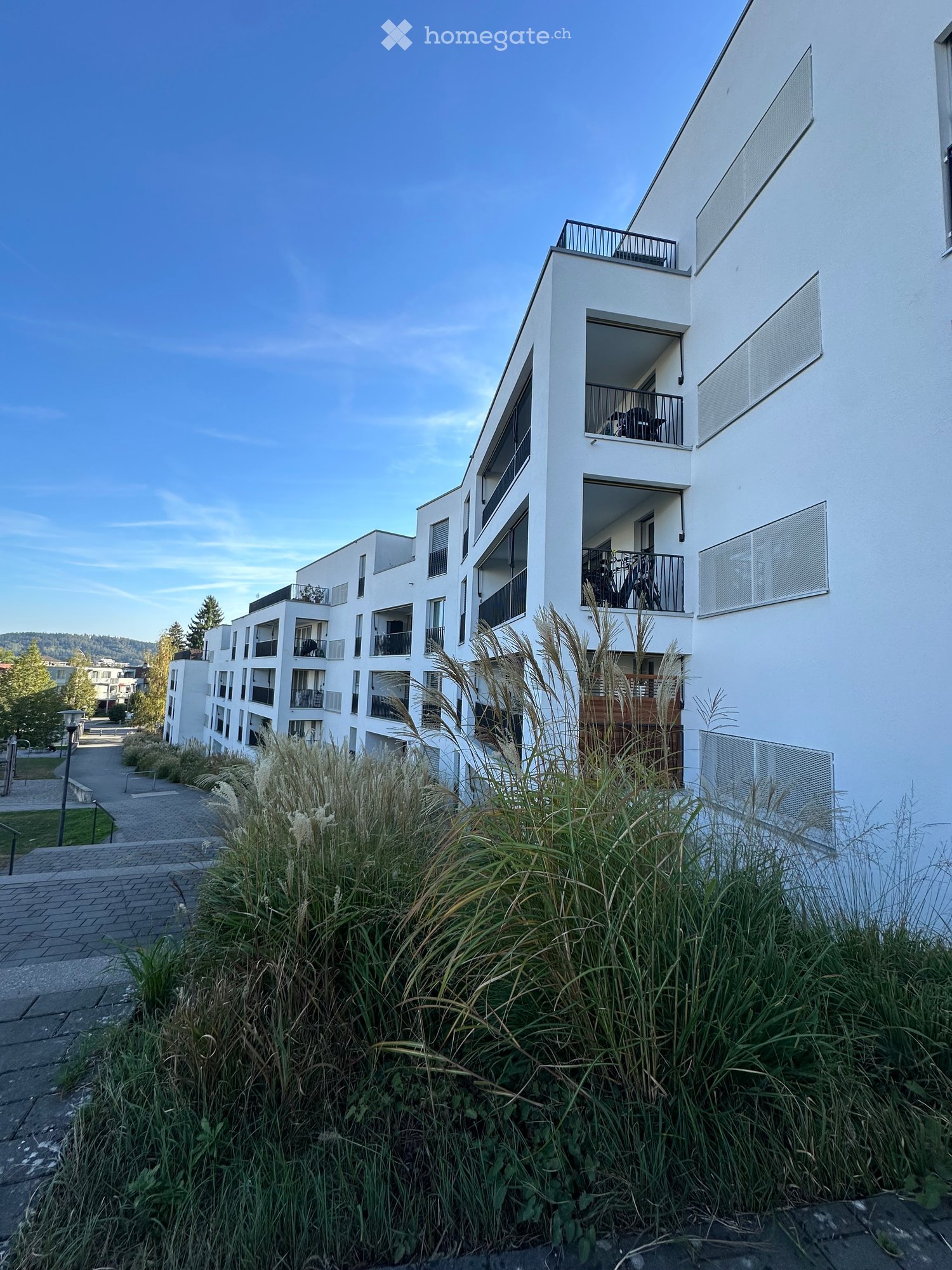 Modern multi-story residential building with white exterior, balconies, and greenery in front.
