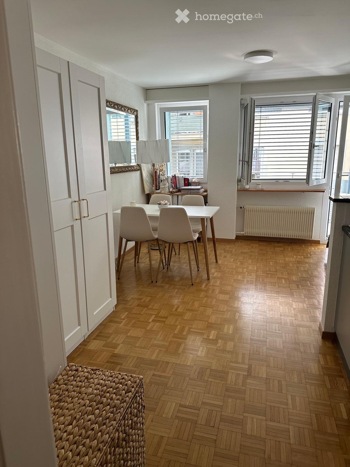 Dining area with wooden herringbone flooring, white table and chairs, large windows with blinds, radiator, and a woven basket on the floor.