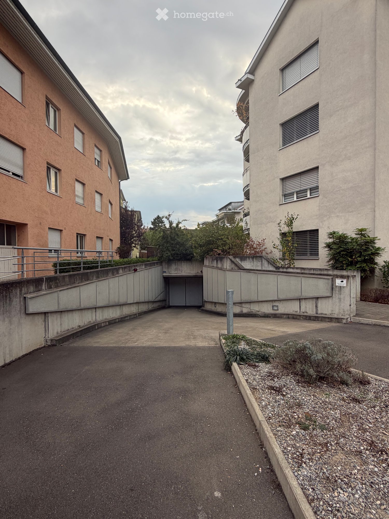 A paved driveway leading to a garage entrance, flanked by modern apartment buildings with balconies and windows.