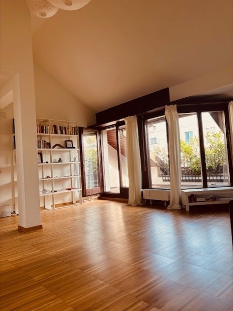 Bright, open-plan room with wooden flooring, large windows, and a balcony. Features a white bookshelf with books and decorative items, a radiator under the windows, and a small table near the balcony door.