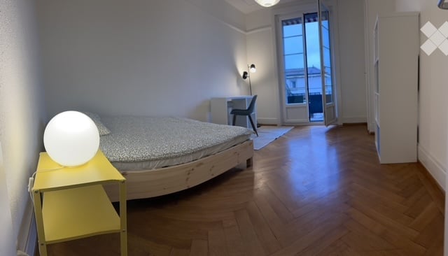 Bright bedroom with wooden flooring, a double bed with patterned bedding, a yellow nightstand with a round lamp, a desk with a chair, and large windows with a view.