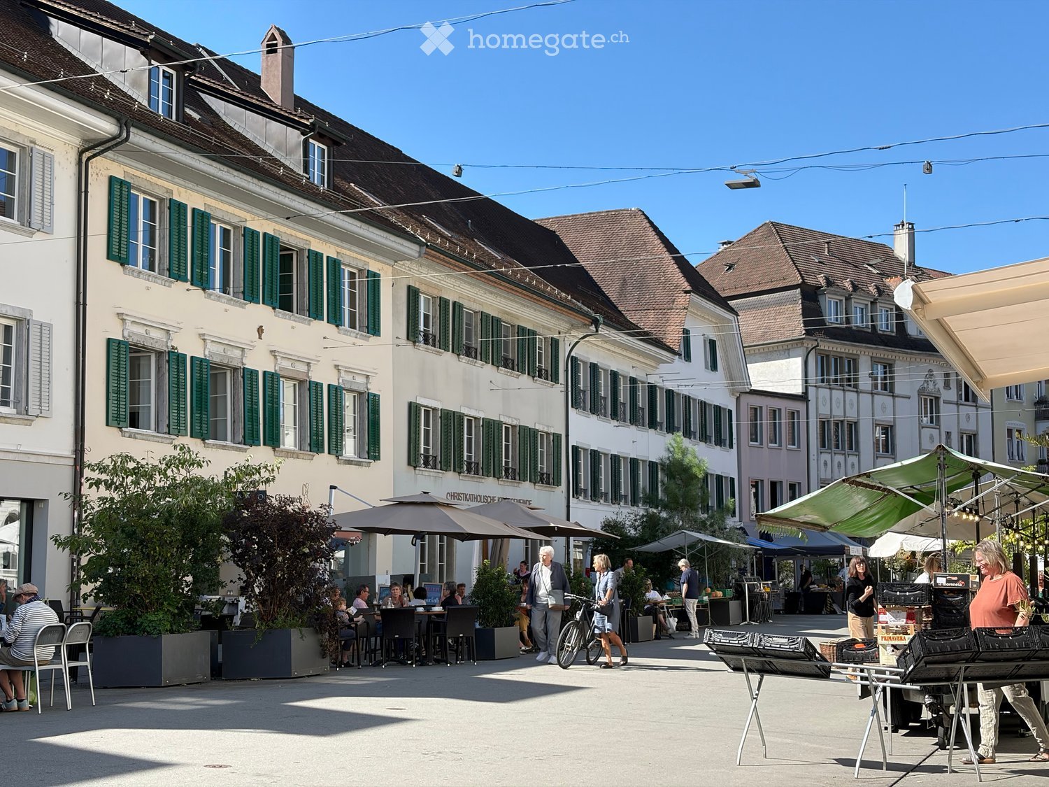 A bustling outdoor area with people sitting at tables under umbrellas, surrounded by buildings with green shutters and a clear blue sky.