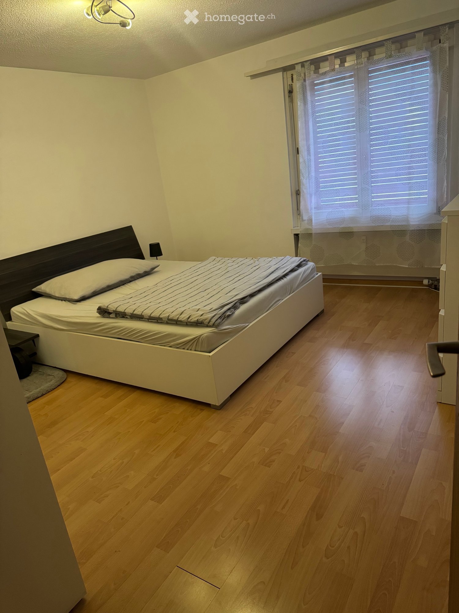 Bedroom with wooden flooring, a white bed with striped bedding, a nightstand with a lamp, and a window with blinds.