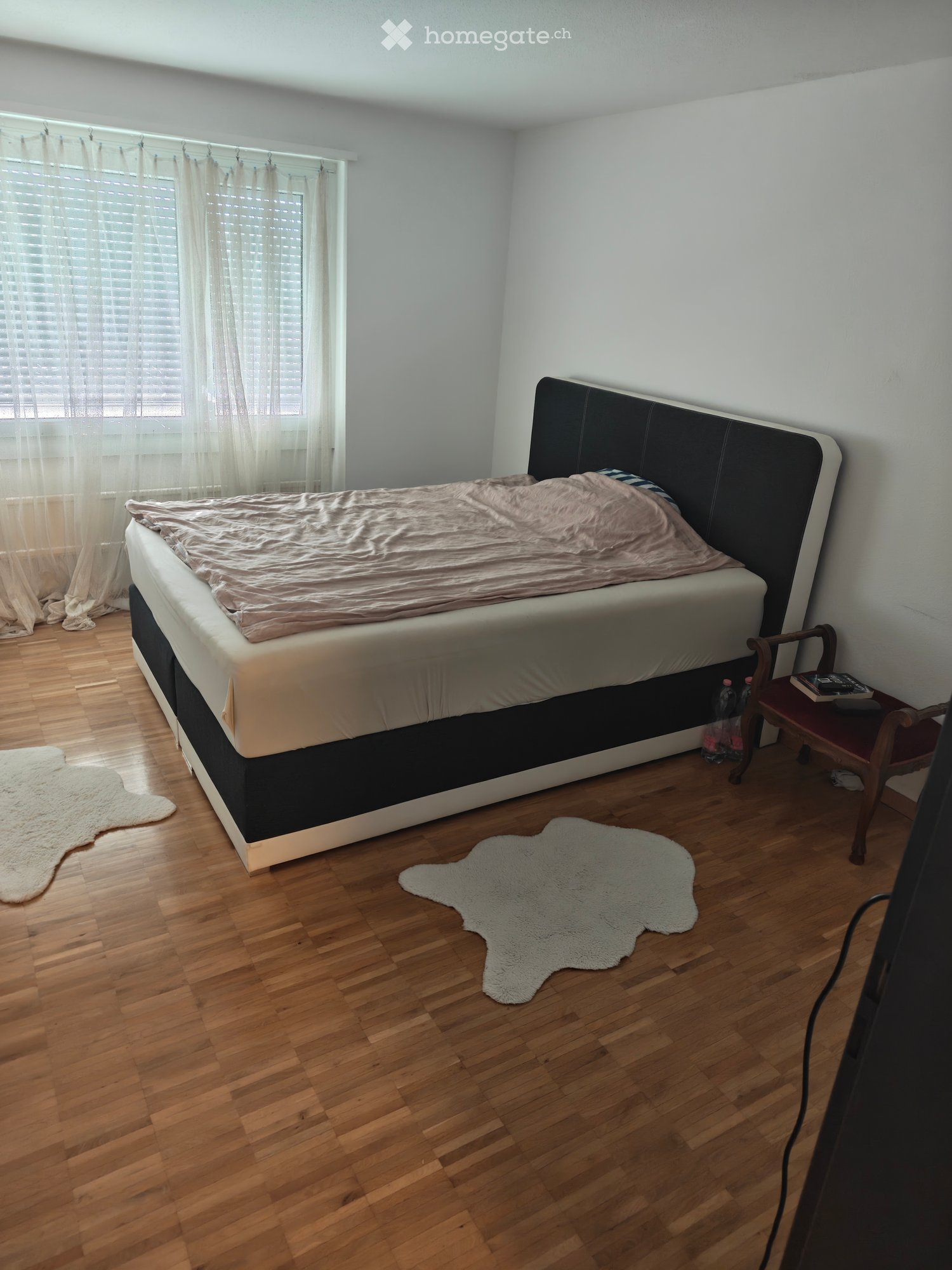 A well-lit bedroom with a large bed, a wooden chair, a small table, and two white rugs on the floor.