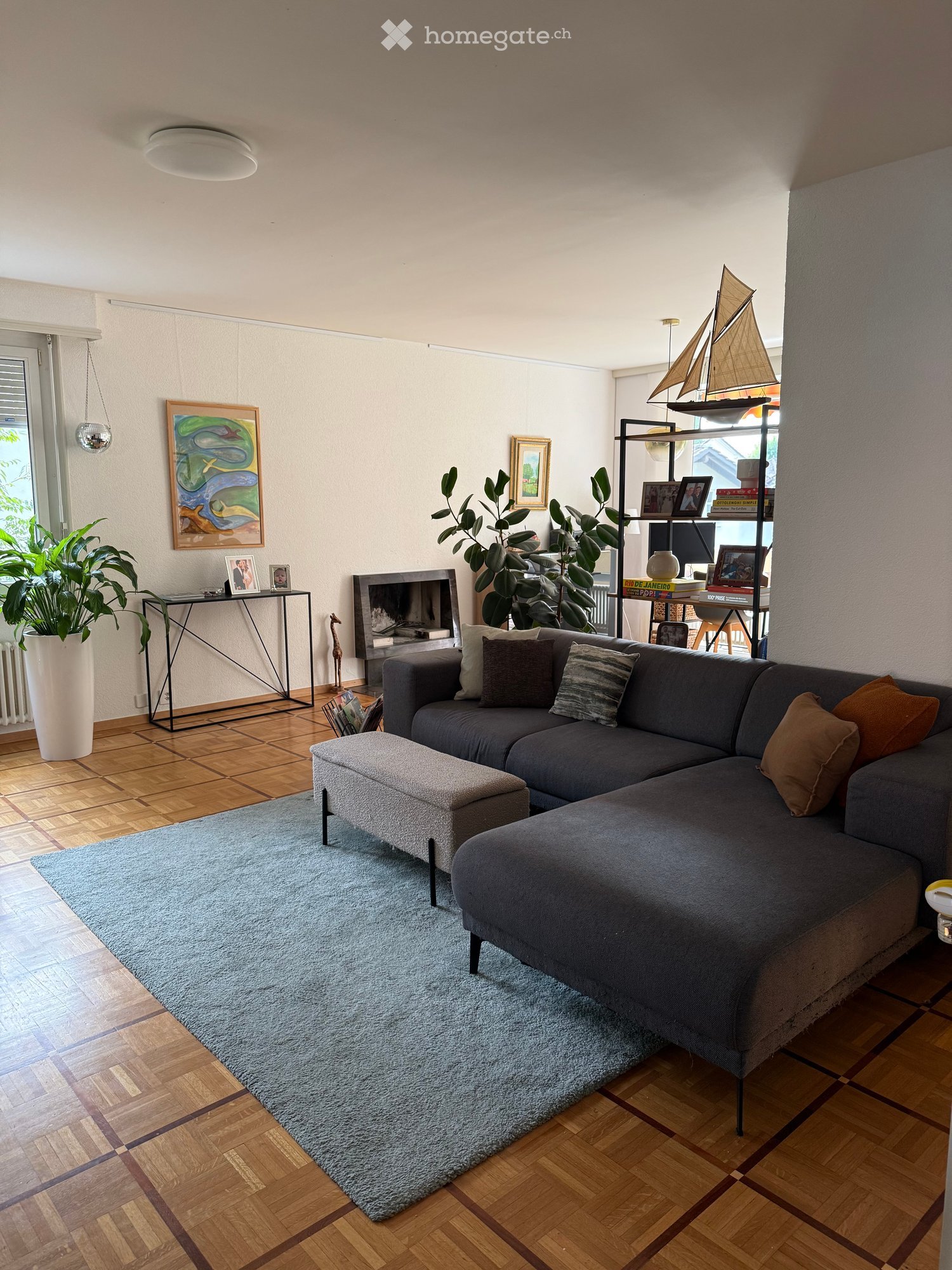 Modern living room with a grey sectional sofa, ottoman, coffee table, fireplace, and decorative shelf with books and a model sailboat.