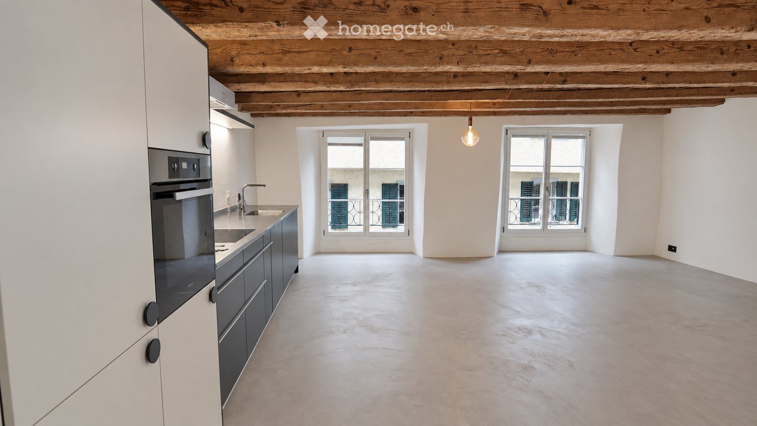 An unfurnished room with a modern kitchen area on the left, featuring white cabinets, a sink, and an oven. The main space is open with concrete flooring, large windows, and exposed wooden beams on the ceiling.