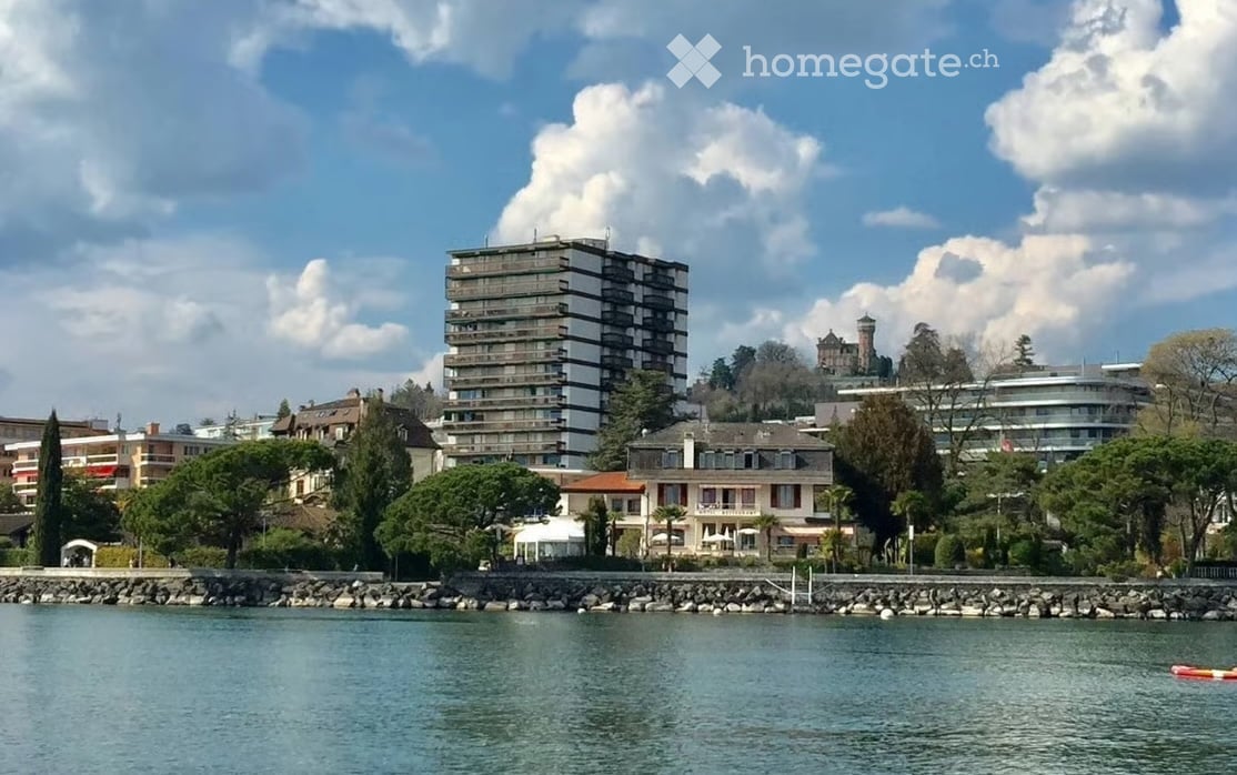 A scenic view of a waterfront area with a mix of residential and commercial buildings, including a prominent high-rise apartment building, a historic castle-like structure, and a variety of smaller houses and trees.