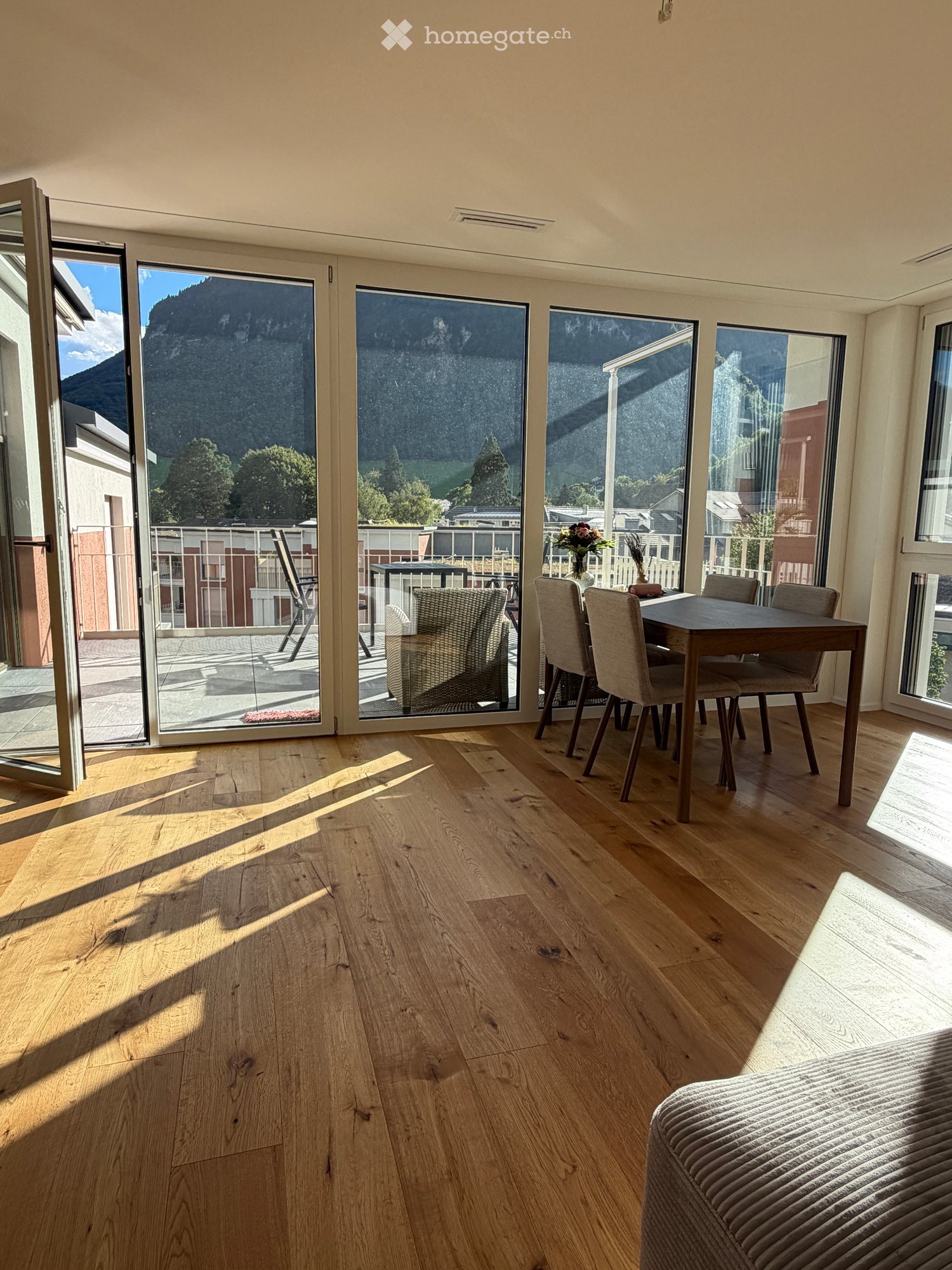 Bright room with wooden flooring, large windows offering a scenic mountain view, a sofa, a dining table with chairs, and a flower vase on the table.