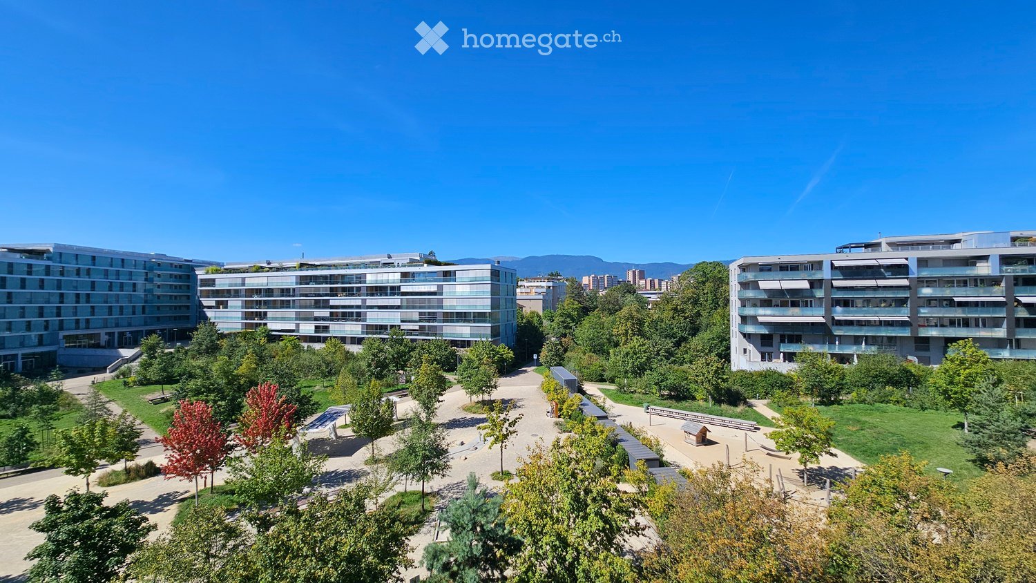 A modern residential complex with contemporary buildings, surrounded by a landscaped park area with pathways, benches, and trees.