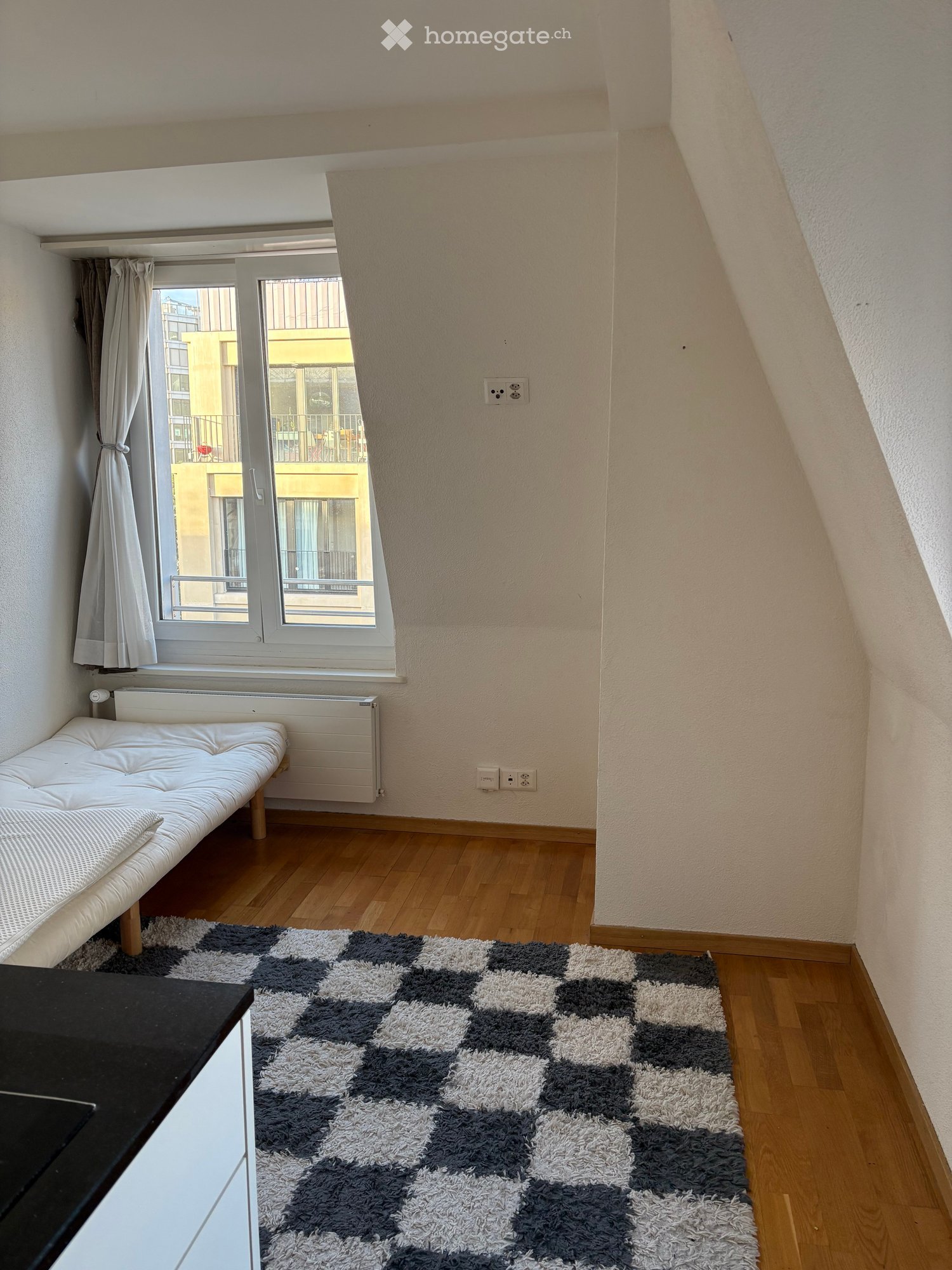 A small, modern room with wooden flooring, a white sofa, a patterned rug, a radiator, and a large window with a curtain. The room has a slanted ceiling typical of an attic space.