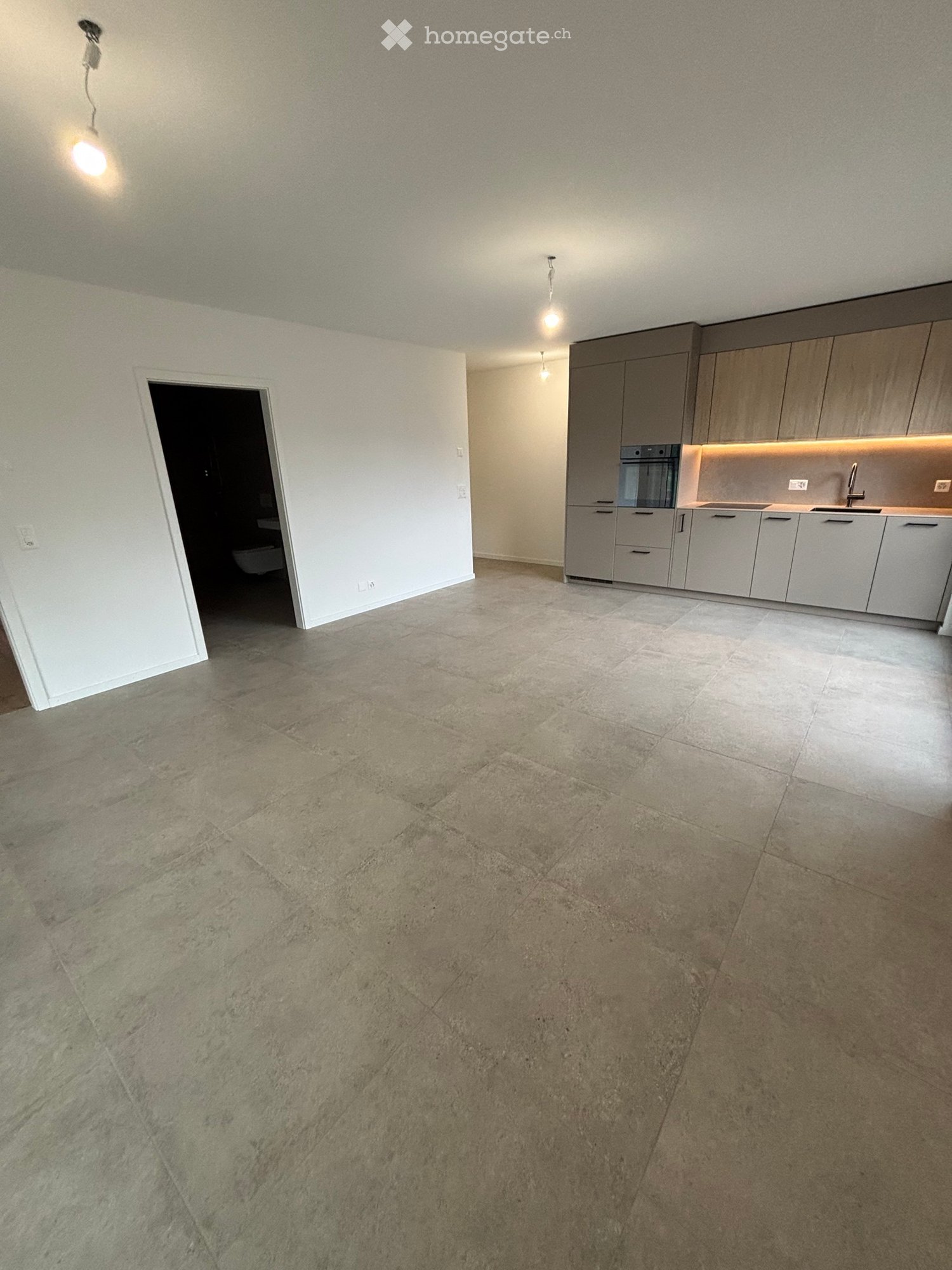Empty room with tiled floor, white walls, and a kitchen area with cabinets and appliances visible in the background.