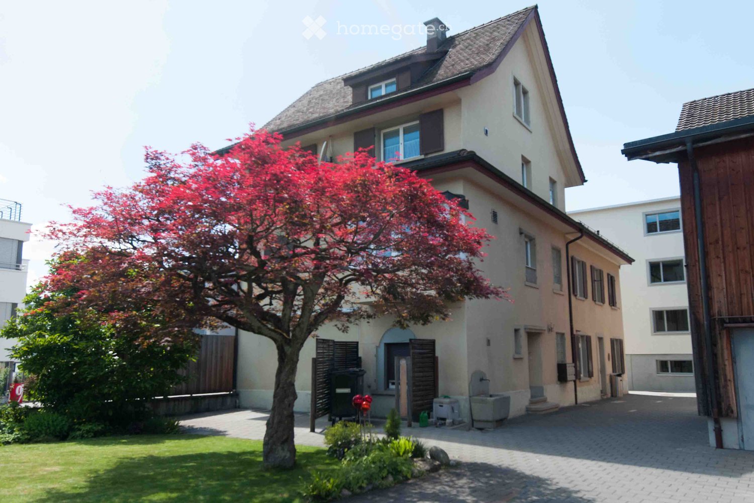 Two-story house with beige walls, multiple windows, and a well-maintained garden with a red-leaved tree and green lawn.