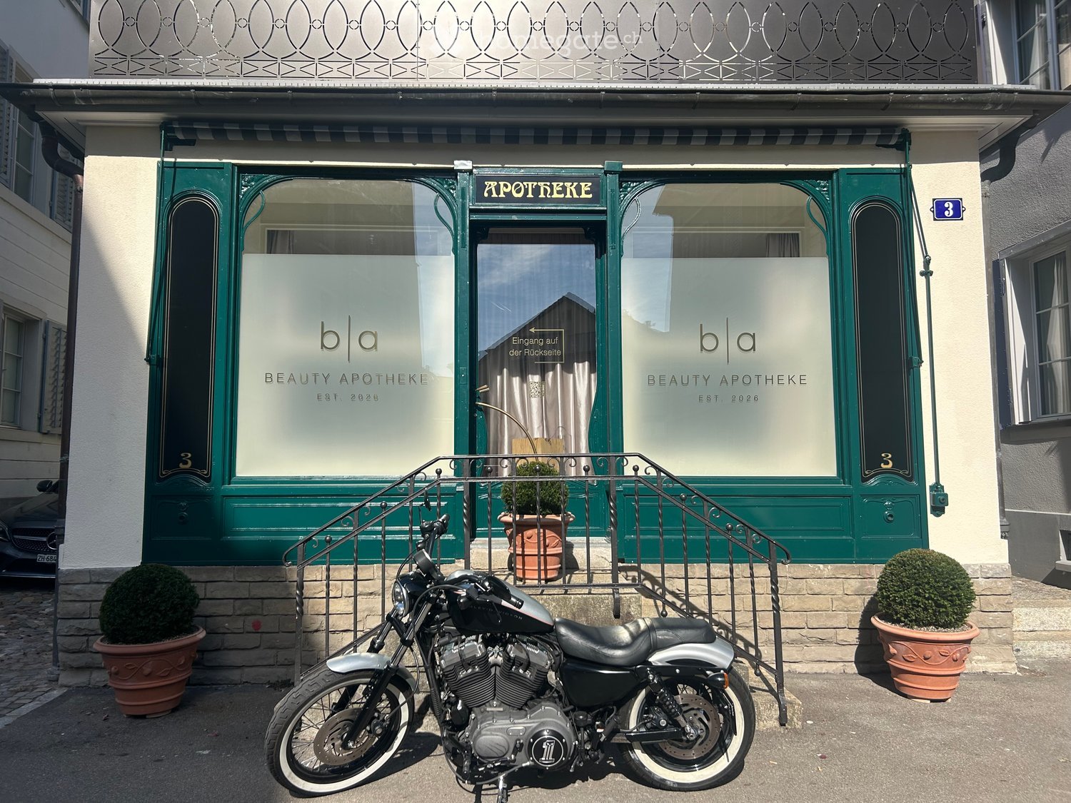 Beauty apotheke storefront with green doors and windows, decorative wrought iron balcony above, potted plants, and a parked motorcycle in front.